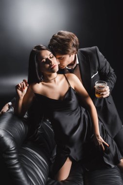 Man in suit holding whiskey and kissing sexy woman in silk dress on couch on black background