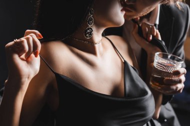 Cropped view of sensual woman in silk dress kissing boyfriend with whiskey isolated on black 
