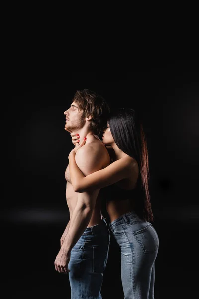 穿着牛仔裤的激情女人在黑色背景下摸着肌肉发达的男朋友的脖子的侧视图 — 图库照片