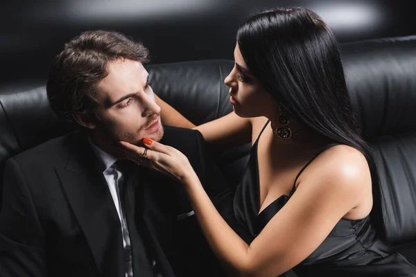 Sexy woman in satin dress touching boyfriend in suit on couch on black background