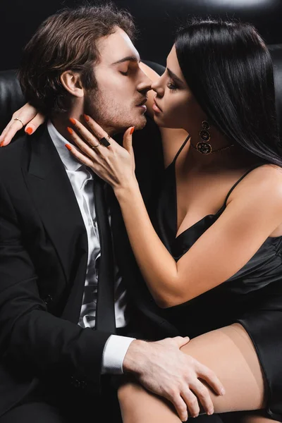 Side view of sexy woman in dress touching boyfriend in suit on couch on black background