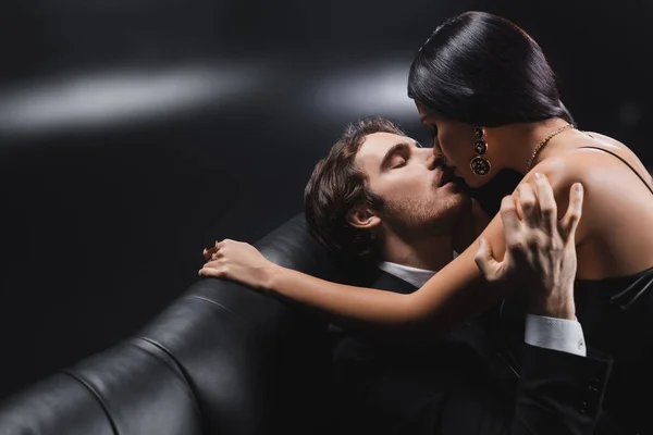 Young man in suit kissing brunette girlfriend on couch on black background