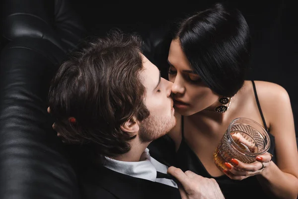 High angle view of brunette woman holding whiskey and kissing boyfriend in suit on couch on black background