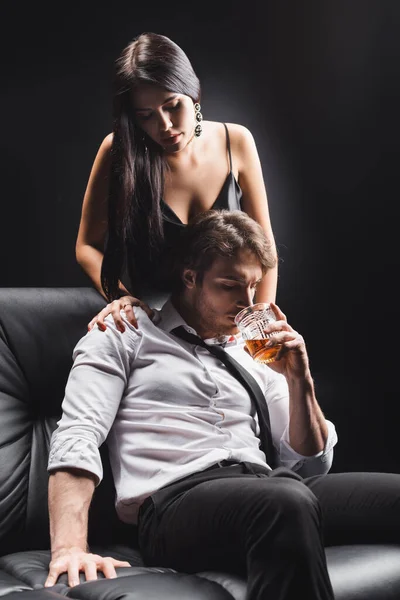 Young man in shirt drinking whiskey near sexy brunette woman isolated on black
