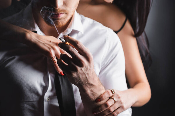 Cropped view of blurred woman touching boyfriend in formal wear with cigarette on black background
