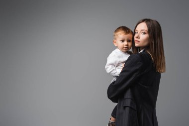 woman in black blazer holding toddler son and looking at camera on grey