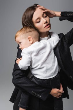 woman with closed eyes touching head while holding baby boy isolated on grey