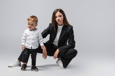 businesswoman in black suit posing near little son standing on grey