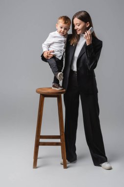happy woman with mobile phone hugging son standing on high stool on grey