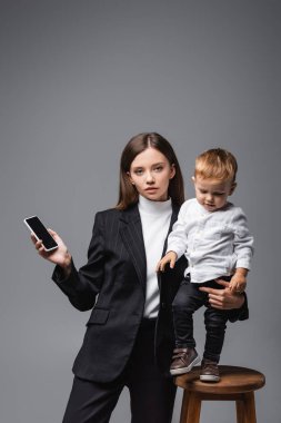 woman holding smartphone with blank screen and toddler son isolated on grey