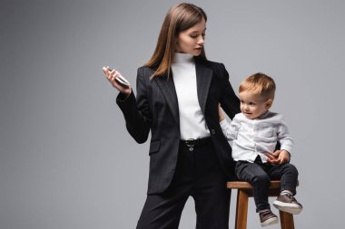 woman in black suit standing with smartphone near son sitting on high stool isolated on grey