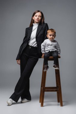 baby boy sitting on high stool and looking at camera near mom in black suit on grey