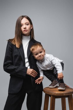 woman in black suit looking at camera near cheerful son grimacing isolated on grey