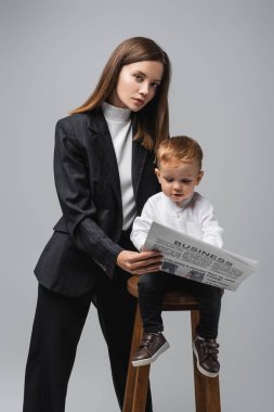 woman in black suit holding business newspaper near toddler son isolated on grey