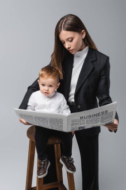 businesswoman reading newspaper near toddler son on high stool isolated on grey