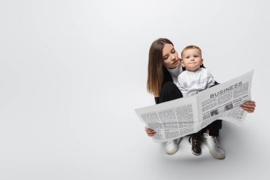 young woman reading newspaper near smiling son on grey