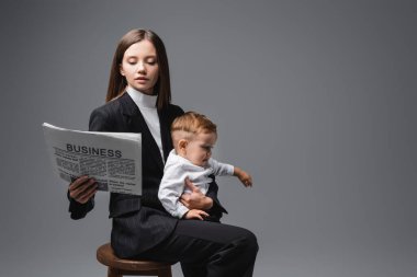 woman in black suit reading business newspaper while sitting with son on high stool isolated on grey