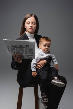 woman reading business newspaper while sitting with little son isolated on grey