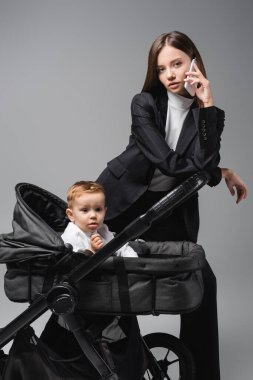 woman in suit talking on mobile phone near toddler son in baby carriage isolated on grey clipart