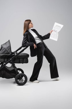 full length view of woman reading newspaper while leaning on black pram on grey clipart