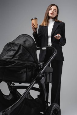 woman in suit holding smartphone and paper cup near black pram isolated on grey clipart