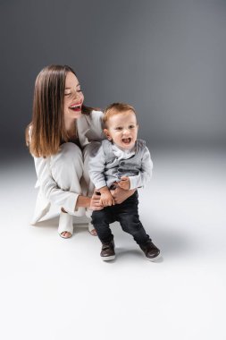 Mutlu bir kadın, şaşkın oğlunu kucaklıyor ve gri üzerinde ağzı açık duruyor.