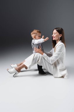 Bebek çocuk, uzanmış elleriyle mutlu annesinin yanında gri üzerinde oturuyor.