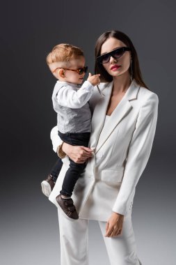 trendy woman in sunglasses holding little son and looking at camera on grey