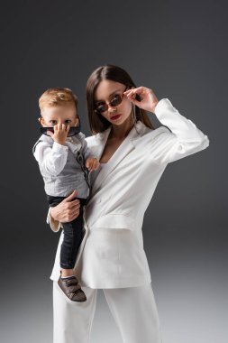 trendy mother and son adjusting sunglasses while looking at camera on grey