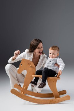 excited woman showing win gesture near son riding rocking horse on grey