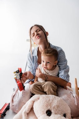 tired woman with closed eyes embracing son and holding toy train on white