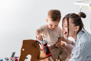 little boy holding toy train near amazed mother on grey