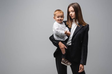 young businesswoman holding little son smiling at camera isolated on grey