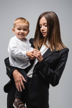 young woman in black suit holding hands of happy son isolated on grey
