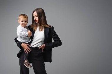 businesswoman standing with hand on hip while holding smiling son isolated on grey
