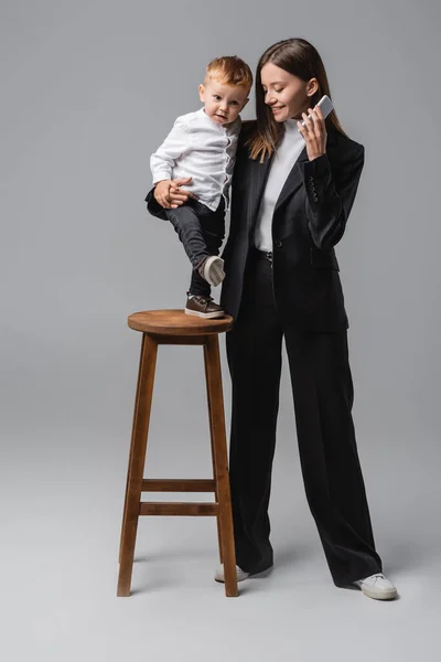 Happy Woman Mobile Phone Hugging Son Standing High Stool Grey — Foto de Stock