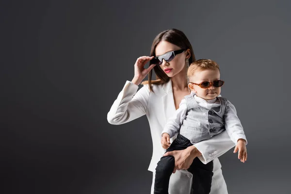 Young Woman Adjusting Stylish Sunglasses While Holding Son Isolated Grey — Foto de Stock