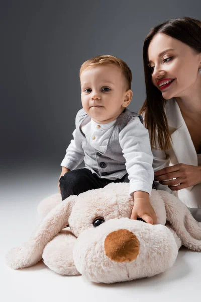 little boy looking at camera while sitting on huge toy dog near smiling mom on grey