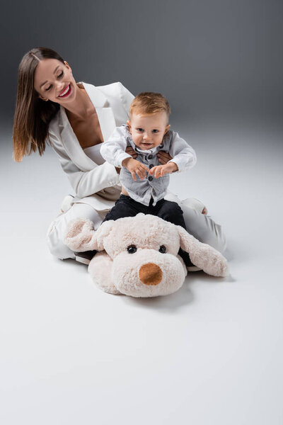 young woman in white suit playing with little kid sitting on toy dog on grey