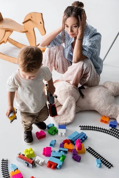 tired woman touching head near son playing with toys on white