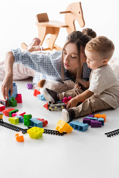 young woman and toddler boy playing with construction cubes and toy railway on white