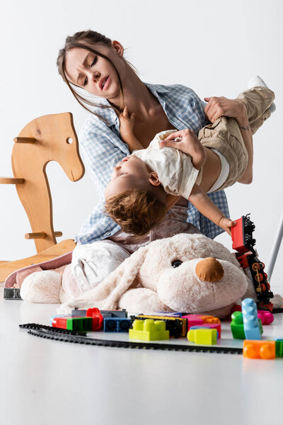 tired woman holding son while playing with him near toys on white