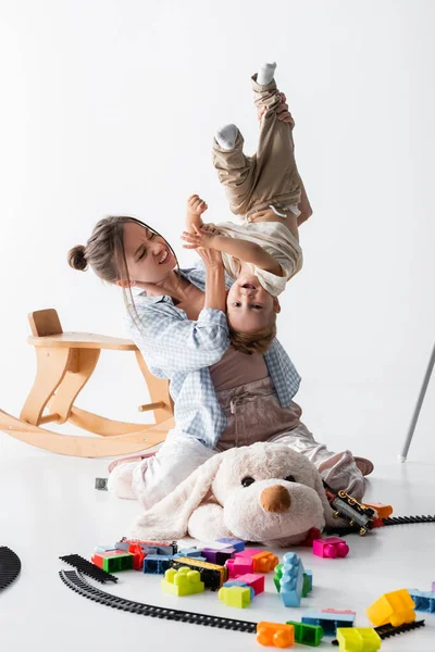 irritated woman holding cheerful son upside down near toys on white