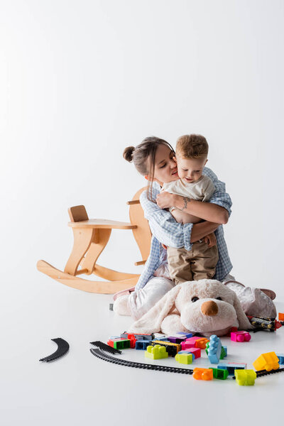 excited woman hugging son near toys and rocking horse on white