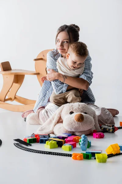 Cansado Mujer Abrazando Alegre Hijo Cerca Juguetes Blanco — Foto de Stock