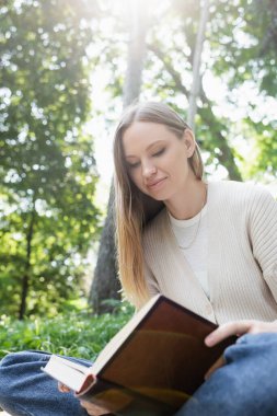 Gülümserken ve yeşil parkta otururken elinde kitap ve gözlük tutan kadın.