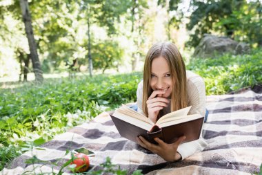 happy woman reading book while lying near smartphone and apple on blanket 