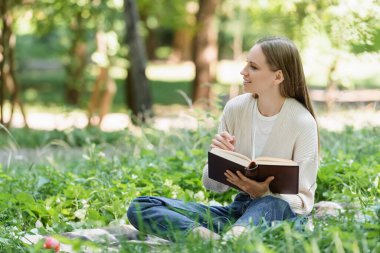 Yeşil çimenlerin üzerinde battaniyeye oturmuş kitabı tutan neşeli bir kadın.
