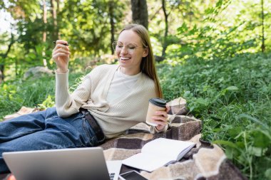 happy woman in glasses holding paper cup and laughing near laptop on blanket in park