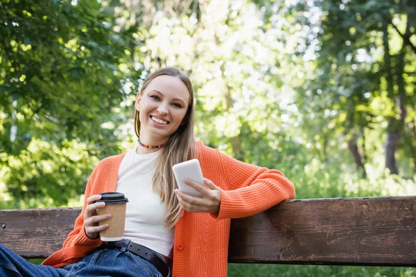 Happy Woman Woman Holding Paper Cup Smartphone While Sitting Bench — Stockfoto